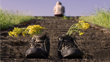 Bildet viser et menneske som sitter med ryggen til øverst i en trapp ute i det fri. Det står et par sko for seg selv på trappen med blomster i.