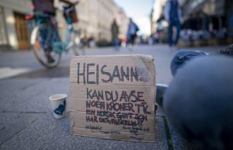 Bildet viser Karl Johans gate og en plakat som en sittende mann har foran seg, hvor det er bønn om bidrag til en som har det vanskelig.