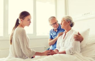 Bildet viser en eldre, syk dame i senga. Familien sitter på sengekanten, og en mann holder rundt henne.