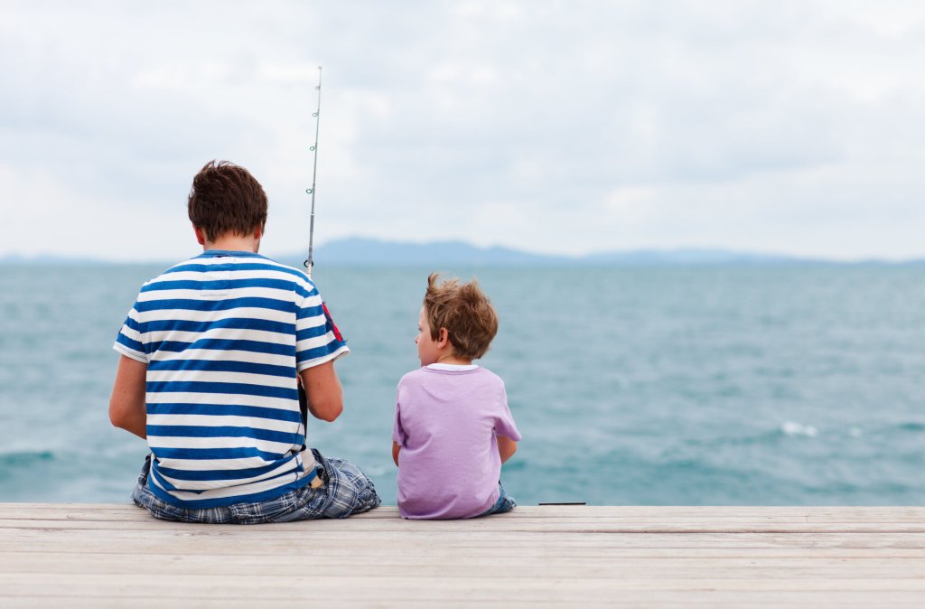 Et lite barn sitter sammen med en voksen mann på en brygge. Mannen fisker.