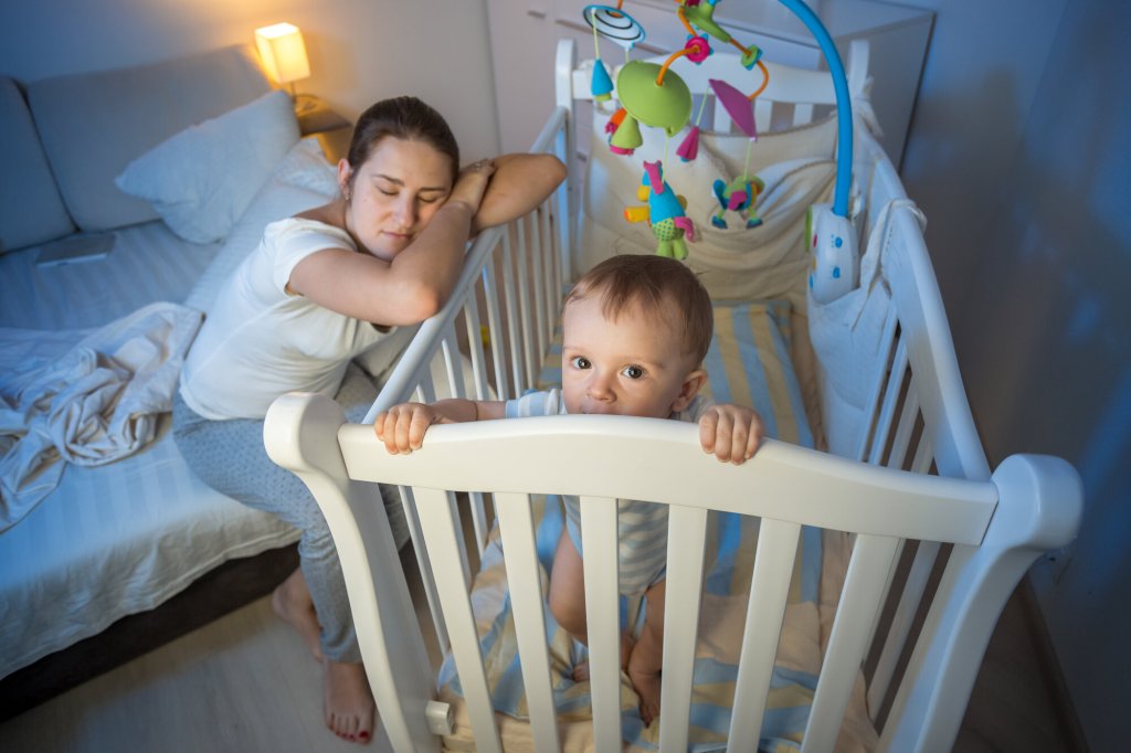 Bildet viser en mor som halvsover over barnesenga, mens barnet står i senga, lys våkent