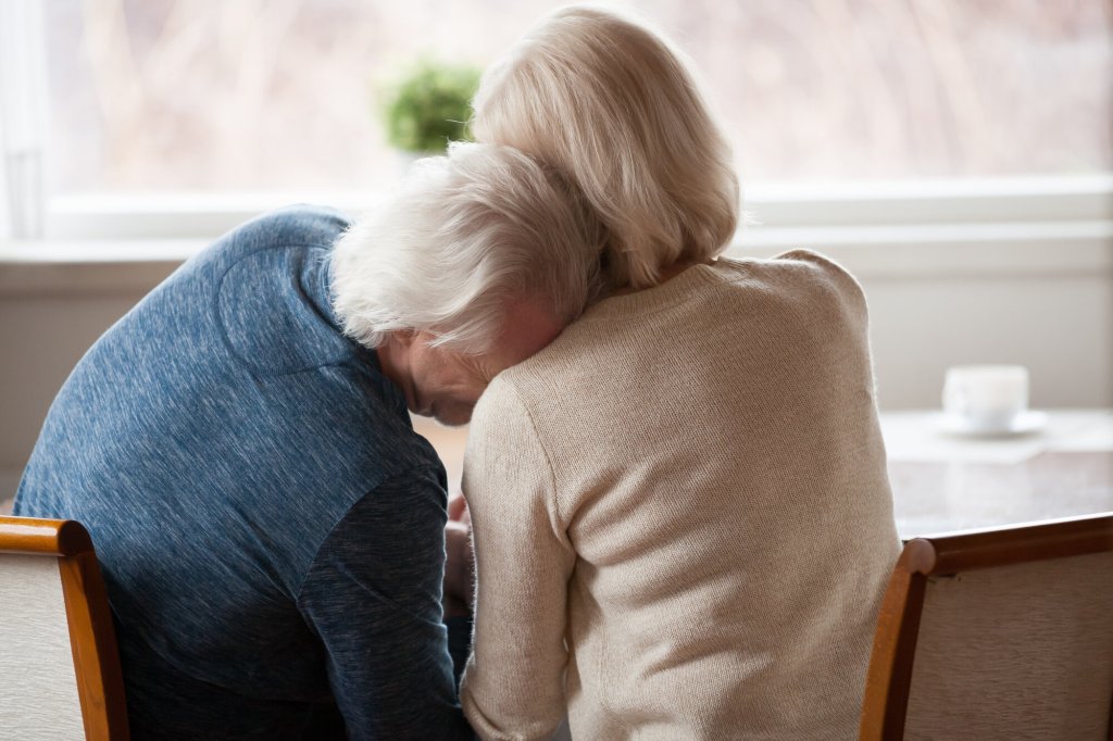 Bildet viser et eldre par som sitter inntil et bord. Mannen lener hodet sitt på kvinnens skulder.