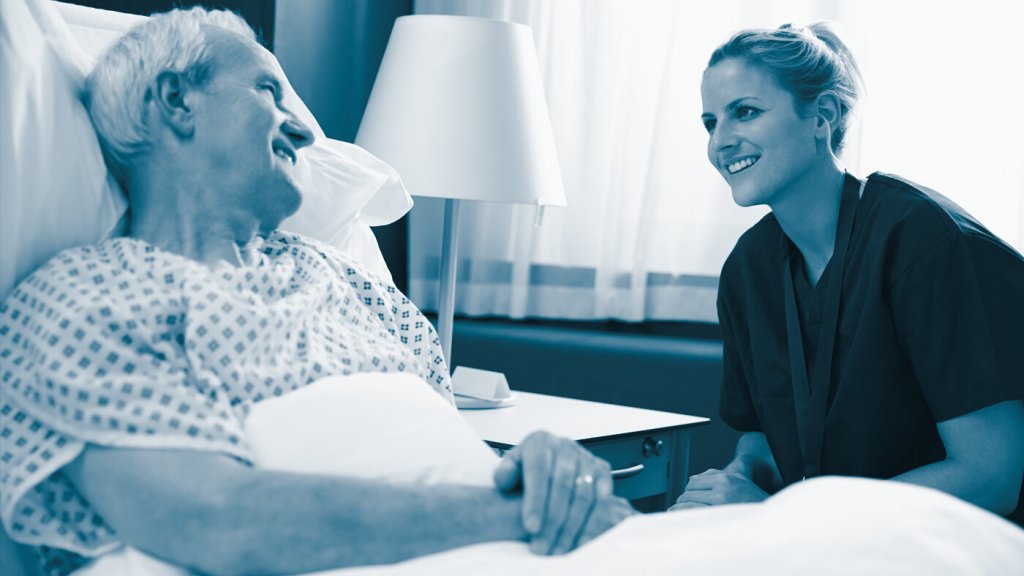 The photo shows a male older patient lying in a hospital bed. A female nurse is sitting beside him. Both are smiling