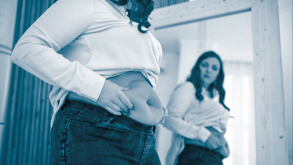 The photo shows a woman looking in the mirror. We can see her belly, and she is squeezing it to show she has a couple of kilos extra