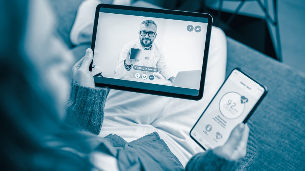 The photo shows a woman sitting in a sofa talking to a doctor via her Ipad. In the other hand she is holding a mobile phone where she is sending her vital data via an app