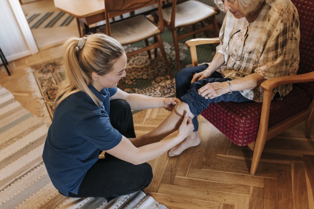 Bildet viser en hjemmesykepleier som skifter strømper på en eldre dame som sitter i stolen i hjemmet sitt.