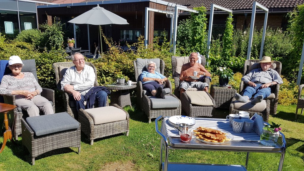 Bildet viser sykehjemsbeboere som sitter i solstoler i hagen og nyter solen. Foran står et bord med vafler.